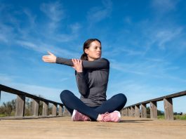 Frau sitzt nach dem Sport auf einer Holzbrücke im Freien und dehnt sich an der Schulter