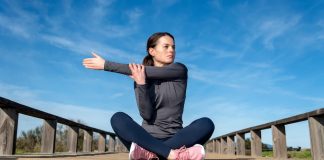 Frau sitzt nach dem Sport auf einer Holzbrücke im Freien und dehnt sich an der Schulter