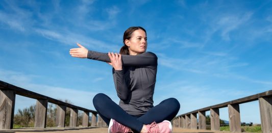Schultergürtel verspannt trotz Bewegung Frau sitzt nach dem Sport auf einer Holzbrücke im Freien und dehnt sich an der Schulter